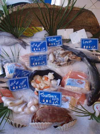 A display of various seafood items on ice, including whole fish, shellfish, and packaged items, with price tags in French. The arrangement is decorated with green palm-like leaves.