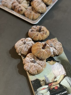 A collection of round bread rolls, dusted with powdered sugar, arranged on a dark surface. The bread is pumpkin-shaped, possibly with raisins or chocolate chips baked inside. In the background, a tray with more of the same bread is partially visible. A book featuring an illustration of a woman in a kitchen setting, possibly cooking, is placed beneath some of the bread rolls.