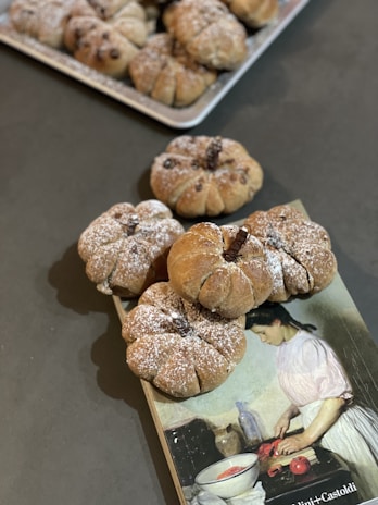 A collection of round bread rolls, dusted with powdered sugar, arranged on a dark surface. The bread is pumpkin-shaped, possibly with raisins or chocolate chips baked inside. In the background, a tray with more of the same bread is partially visible. A book featuring an illustration of a woman in a kitchen setting, possibly cooking, is placed beneath some of the bread rolls.