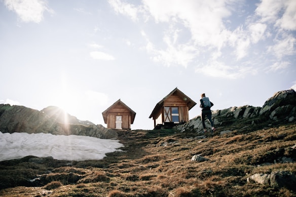 Two small wooden cabins are situated on a hillside with a person wearing a backpack walking towards them. The sky is mostly clear with a sunbeam peeking through, casting light over the scene. A patch of snow can be seen on the left side among the rocky terrain.