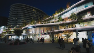 A modern, multi-story shopping complex is bustling with people in the evening. The building features sleek, curved architecture with illuminated signs and greenery on various levels. Trees with autumn leaves and brightly lit shop windows create a vibrant atmosphere.