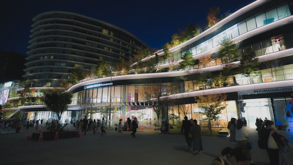 A modern, multi-story shopping complex is bustling with people in the evening. The building features sleek, curved architecture with illuminated signs and greenery on various levels. Trees with autumn leaves and brightly lit shop windows create a vibrant atmosphere.