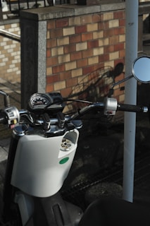 A close-up view of a scooter's handlebars and dashboard, including rearview mirrors and a speedometer. The scooter is parked in front of a brick wall, casting a shadow on the ground. The scene is captured in natural daylight, emphasizing the textures of the scooter and the background.
