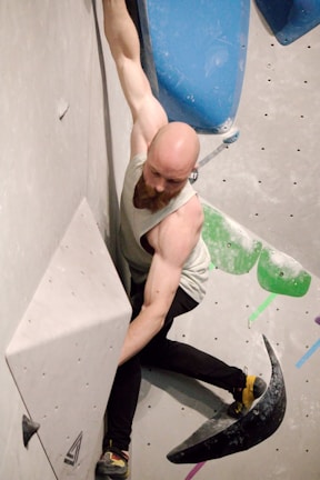 A person is engaging in indoor rock climbing, demonstrating strength and focus while navigating a climbing wall with holds of various shapes and colors. The climber is using both hands and feet, wearing a sleeveless shirt and climbing shoes. The wall displays bold geometric shapes and subtle color variations.