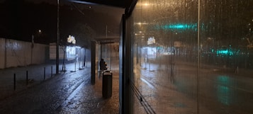 A rainy night scene at a bus stop, with the ground wet and shining with reflections from streetlights. A person wearing a hoodie is sitting alone on a bench, while bright lights from a nearby kiosk glow in the distance. Raindrops are visible on the glass of the bus stop shelter, creating an atmospheric, moody effect.