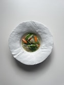A minimalistic and elegant dish is presented in a textured white bowl. The dish features a small portion of vibrant vegetables, including carrots, onions, and green leafy vegetables, resting in a clear broth.