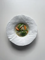 A delicate porcelain bowl holding a clear vegetable broth with colorful diced carrots, celery, and herbs floating gently.