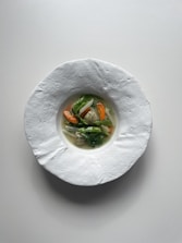 A minimalistic and elegant dish is presented in a textured white bowl. The dish features a small portion of vibrant vegetables, including carrots, onions, and green leafy vegetables, resting in a clear broth.