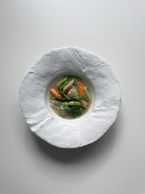 A minimalistic and elegant dish is presented in a textured white bowl. The dish features a small portion of vibrant vegetables, including carrots, onions, and green leafy vegetables, resting in a clear broth.