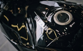 A close-up view of a shiny black motorcycle fuel tank with the brand name 'Suzuki' prominently displayed. The fuel cap is visible on the right side, surrounded by a circular metallic fixture. The surface reflects light and surroundings, creating an abstract pattern with glossy highlights.