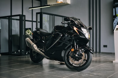 A sleek black motorcycle with a shiny exterior is positioned in a modern showroom environment. The motorcycle features aerodynamic design elements with prominent front headlights and dual exhaust pipes. The floor is made of glossy tiles, and the space is separated by glass partitions. The ambiance suggests a premium, sophisticated atmosphere.