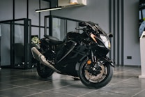 A sleek black motorcycle with a shiny exterior is positioned in a modern showroom environment. The motorcycle features aerodynamic design elements with prominent front headlights and dual exhaust pipes. The floor is made of glossy tiles, and the space is separated by glass partitions. The ambiance suggests a premium, sophisticated atmosphere.