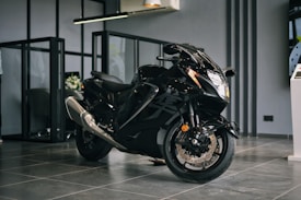 A sleek black motorcycle with a shiny exterior is positioned in a modern showroom environment. The motorcycle features aerodynamic design elements with prominent front headlights and dual exhaust pipes. The floor is made of glossy tiles, and the space is separated by glass partitions. The ambiance suggests a premium, sophisticated atmosphere.