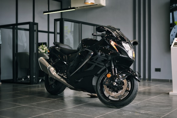 A sleek black motorcycle with a shiny exterior is positioned in a modern showroom environment. The motorcycle features aerodynamic design elements with prominent front headlights and dual exhaust pipes. The floor is made of glossy tiles, and the space is separated by glass partitions. The ambiance suggests a premium, sophisticated atmosphere.