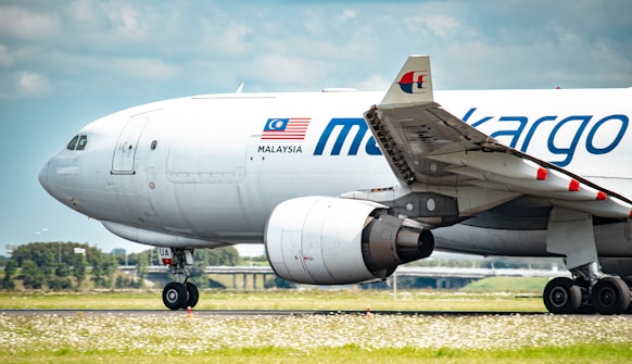 A cargo airplane with 'MALAYSIA' and 'masked off text'-branded livery is taxiing on a runway. The aircraft is predominantly white with red, blue, and gray detailing. The Malaysian flag is visible on the fuselage. The runway area features grass and flowers, and the sky is partly cloudy.