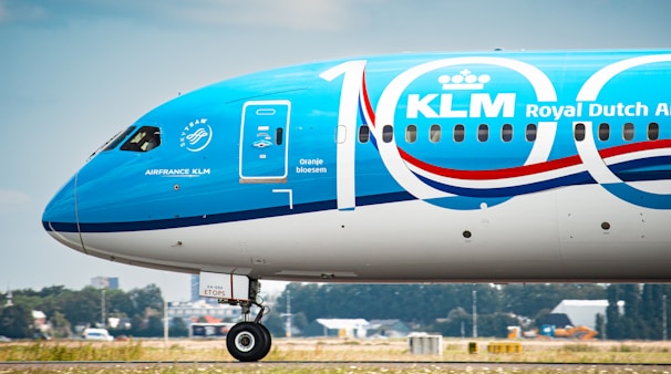 A close-up view of a KLM Royal Dutch Airlines airplane with a special 100-year anniversary livery. The aircraft is blue and white with large '100' numerals prominently displayed. The SkyTeam logo and KLM name are visible. The aircraft is on the runway with a blurry landscape in the background.