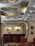 A stylish interior of a café or restaurant featuring elegant, circular ceiling lights in a retro design. The room is furnished with red velvet chairs and booths that provide a warm and inviting atmosphere. Framed artwork and a coat rack are visible on the walls, adding to the cozy ambiance. The color palette includes deep reds and soft creams, with wooden accents.