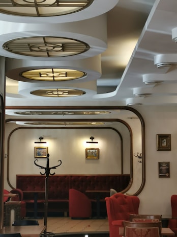A stylish interior of a café or restaurant featuring elegant, circular ceiling lights in a retro design. The room is furnished with red velvet chairs and booths that provide a warm and inviting atmosphere. Framed artwork and a coat rack are visible on the walls, adding to the cozy ambiance. The color palette includes deep reds and soft creams, with wooden accents.
