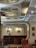 A stylish interior of a café or restaurant featuring elegant, circular ceiling lights in a retro design. The room is furnished with red velvet chairs and booths that provide a warm and inviting atmosphere. Framed artwork and a coat rack are visible on the walls, adding to the cozy ambiance. The color palette includes deep reds and soft creams, with wooden accents.