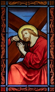 A stained glass artwork depicting a religious figure, shown with a halo around his head, carrying a large wooden cross. He is dressed in red robes and has a solemn expression on his face. The background consists of blue and brown segments, framed with intricate designs in red and gold.
