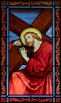 A stained glass artwork depicting a religious figure, shown with a halo around his head, carrying a large wooden cross. He is dressed in red robes and has a solemn expression on his face. The background consists of blue and brown segments, framed with intricate designs in red and gold.