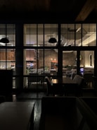 The image depicts the interior of a cozy restaurant, viewed through a grid of large windows. Inside, tables and chairs are neatly arranged, with soft lighting creating a warm atmosphere. A person is seated at a table using a laptop, and several plant decorations add a touch of greenery. The lighting fixtures and wall textures contribute to a modern yet inviting design.