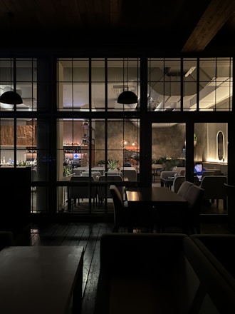 The image depicts the interior of a cozy restaurant, viewed through a grid of large windows. Inside, tables and chairs are neatly arranged, with soft lighting creating a warm atmosphere. A person is seated at a table using a laptop, and several plant decorations add a touch of greenery. The lighting fixtures and wall textures contribute to a modern yet inviting design.
