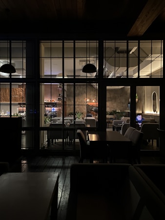 The image depicts the interior of a cozy restaurant, viewed through a grid of large windows. Inside, tables and chairs are neatly arranged, with soft lighting creating a warm atmosphere. A person is seated at a table using a laptop, and several plant decorations add a touch of greenery. The lighting fixtures and wall textures contribute to a modern yet inviting design.