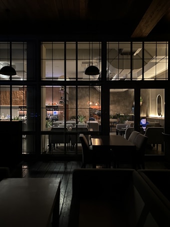 The image depicts the interior of a cozy restaurant, viewed through a grid of large windows. Inside, tables and chairs are neatly arranged, with soft lighting creating a warm atmosphere. A person is seated at a table using a laptop, and several plant decorations add a touch of greenery. The lighting fixtures and wall textures contribute to a modern yet inviting design.