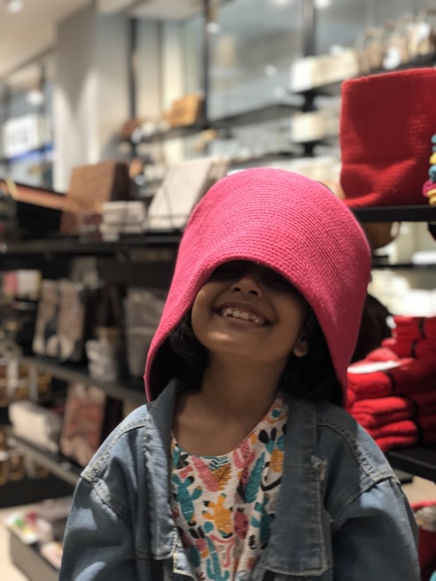 A child is smiling with a pink hat covering their eyes, wearing a denim jacket and a colorful patterned shirt, in a store setting with shelves filled with various items.