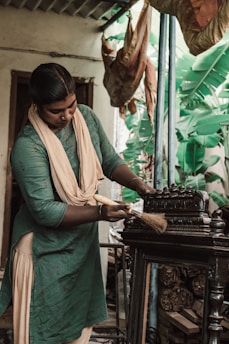 A person is engaged in cleaning or polishing an intricately carved piece of furniture. They are wearing a green and beige outfit and holding a brush in hand. The background includes green banana leaves and drying leaves hanging from the ceiling, suggesting a rustic or traditional setting.