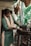 A person is engaged in cleaning or polishing an intricately carved piece of furniture. They are wearing a green and beige outfit and holding a brush in hand. The background includes green banana leaves and drying leaves hanging from the ceiling, suggesting a rustic or traditional setting.