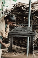 A person wearing traditional clothing is working meticulously on what appears to be an intricately designed wooden furniture piece or artifact. The background shows a large quantity of stacked wooden planks, indicating a workshop or carpentry setting.