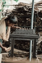 A person wearing traditional clothing is working meticulously on what appears to be an intricately designed wooden furniture piece or artifact. The background shows a large quantity of stacked wooden planks, indicating a workshop or carpentry setting.