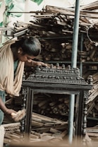 A person wearing traditional clothing is working meticulously on what appears to be an intricately designed wooden furniture piece or artifact. The background shows a large quantity of stacked wooden planks, indicating a workshop or carpentry setting.