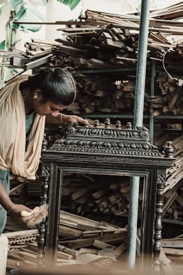 A person wearing traditional clothing is working meticulously on what appears to be an intricately designed wooden furniture piece or artifact. The background shows a large quantity of stacked wooden planks, indicating a workshop or carpentry setting.