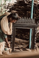 A woman is intensely focused on polishing a large wooden frame. She is wearing traditional clothing and appears to be in a workshop environment. The background is filled with stacks of wooden planks and tools, suggesting a busy woodworking or carpentry space.