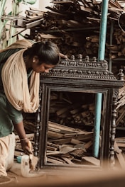 A woman is intensely focused on polishing a large wooden frame. She is wearing traditional clothing and appears to be in a workshop environment. The background is filled with stacks of wooden planks and tools, suggesting a busy woodworking or carpentry space.