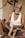 An elderly man with gray hair and a beard is seated on the floor, holding a wooden frame in his hands. He wears a white sleeveless shirt and dark shorts. The background includes a wooden surface and a section of a wall with visible electrical components.
