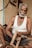 An elderly man with gray hair and a beard is seated on the floor, holding a wooden frame in his hands. He wears a white sleeveless shirt and dark shorts. The background includes a wooden surface and a section of a wall with visible electrical components.