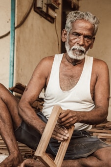 An elderly man with gray hair and a beard is seated on the floor, holding a wooden frame in his hands. He wears a white sleeveless shirt and dark shorts. The background includes a wooden surface and a section of a wall with visible electrical components.