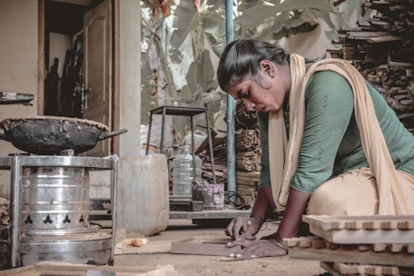 A woman is focused on her work, kneeling on the ground inside a cluttered workshop. She is using her hands to shape or assemble materials. A variety of tools, wooden pieces, and containers are scattered around her. The atmosphere suggests manual labor and craftsmanship.