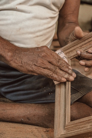 A person is using sandpaper to smooth a wooden frame. The hands and forearms are emphasized, with details of the skin texture visible. The person wears a light-colored shirt and dark shorts, sitting on a surface that has a textured, earthy look.