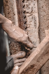 A person is engaged in wood carving, using a chisel to intricately engrave patterns into a piece of wood. The hands are weathered, suggesting experience or age, and the focus is on the detailed craftsmanship. There is a close-up view of tools and the carved wooden surface.