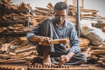 A man is sitting on the ground in a workshop surrounded by stacks of wooden planks. He is focused on his work, holding a piece of wood and a hammer as he constructs or repairs something meticulously. The environment is rustic, filled with wood and tools, suggesting a craft or woodworking setting.