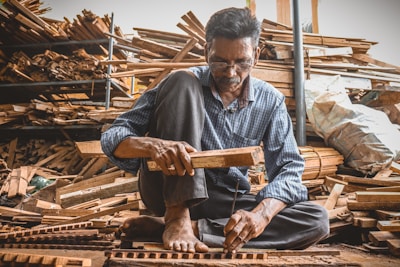 A man is sitting on the ground in a workshop surrounded by stacks of wooden planks. He is focused on his work, holding a piece of wood and a hammer as he constructs or repairs something meticulously. The environment is rustic, filled with wood and tools, suggesting a craft or woodworking setting.