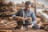 A man is sitting on the ground in a workshop surrounded by stacks of wooden planks. He is focused on his work, holding a piece of wood and a hammer as he constructs or repairs something meticulously. The environment is rustic, filled with wood and tools, suggesting a craft or woodworking setting.