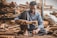 A man is sitting on the ground in a workshop surrounded by stacks of wooden planks. He is focused on his work, holding a piece of wood and a hammer as he constructs or repairs something meticulously. The environment is rustic, filled with wood and tools, suggesting a craft or woodworking setting.