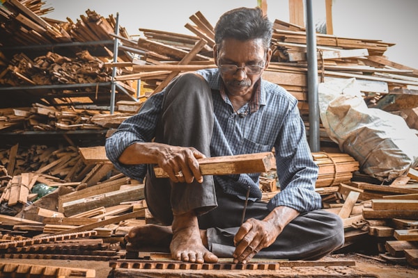 A man is sitting on the ground in a workshop surrounded by stacks of wooden planks. He is focused on his work, holding a piece of wood and a hammer as he constructs or repairs something meticulously. The environment is rustic, filled with wood and tools, suggesting a craft or woodworking setting.