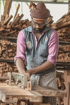 A man is working with a machine in a woodworking shop, wearing a turban and glasses. He is focused on the task at hand, surrounded by neatly stacked pieces of wood. The atmosphere appears to be industrious and traditional, reflecting a wood crafting process.